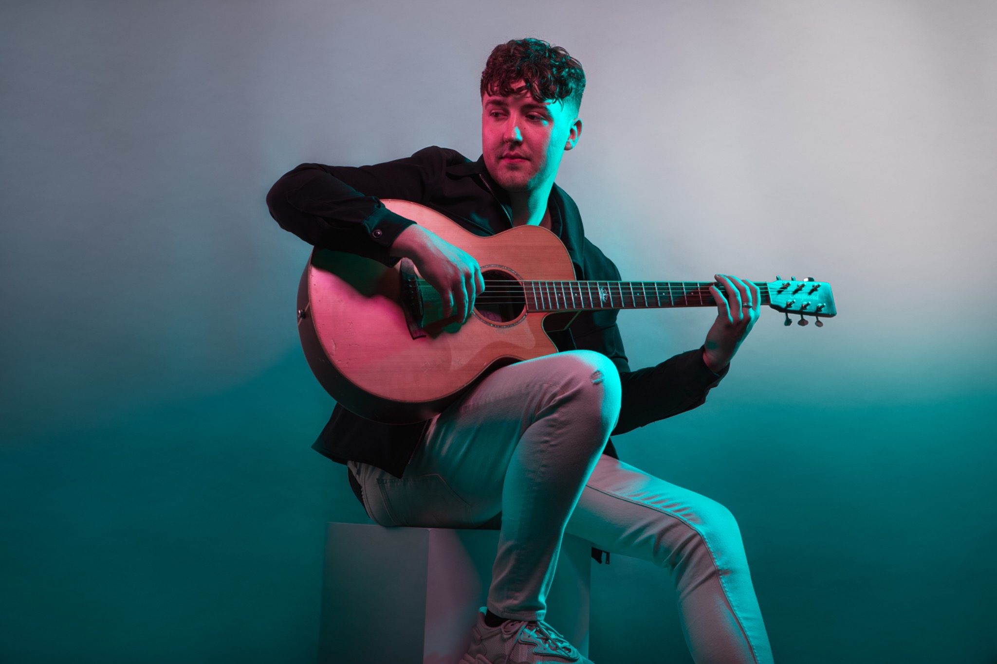 Musician seated with an acoustic guitar under colourful studio lighting